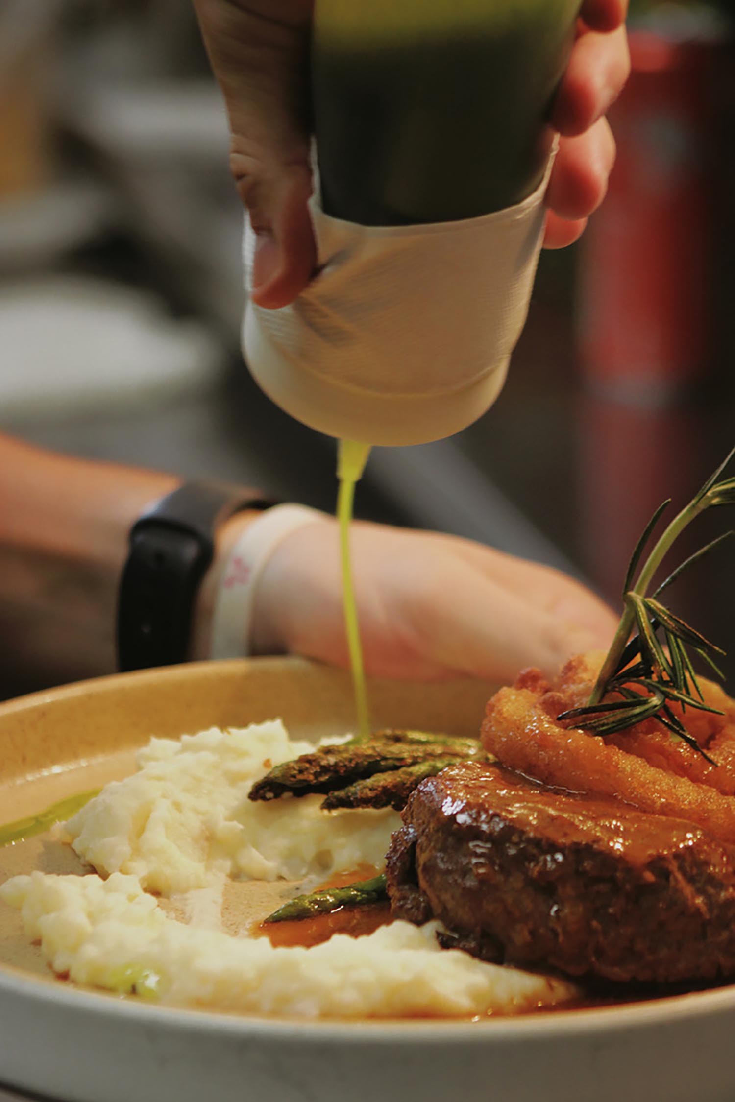 Hand pouring green sauce onto plated steak with mashed potatoes, fried onion rings, and asparagus.