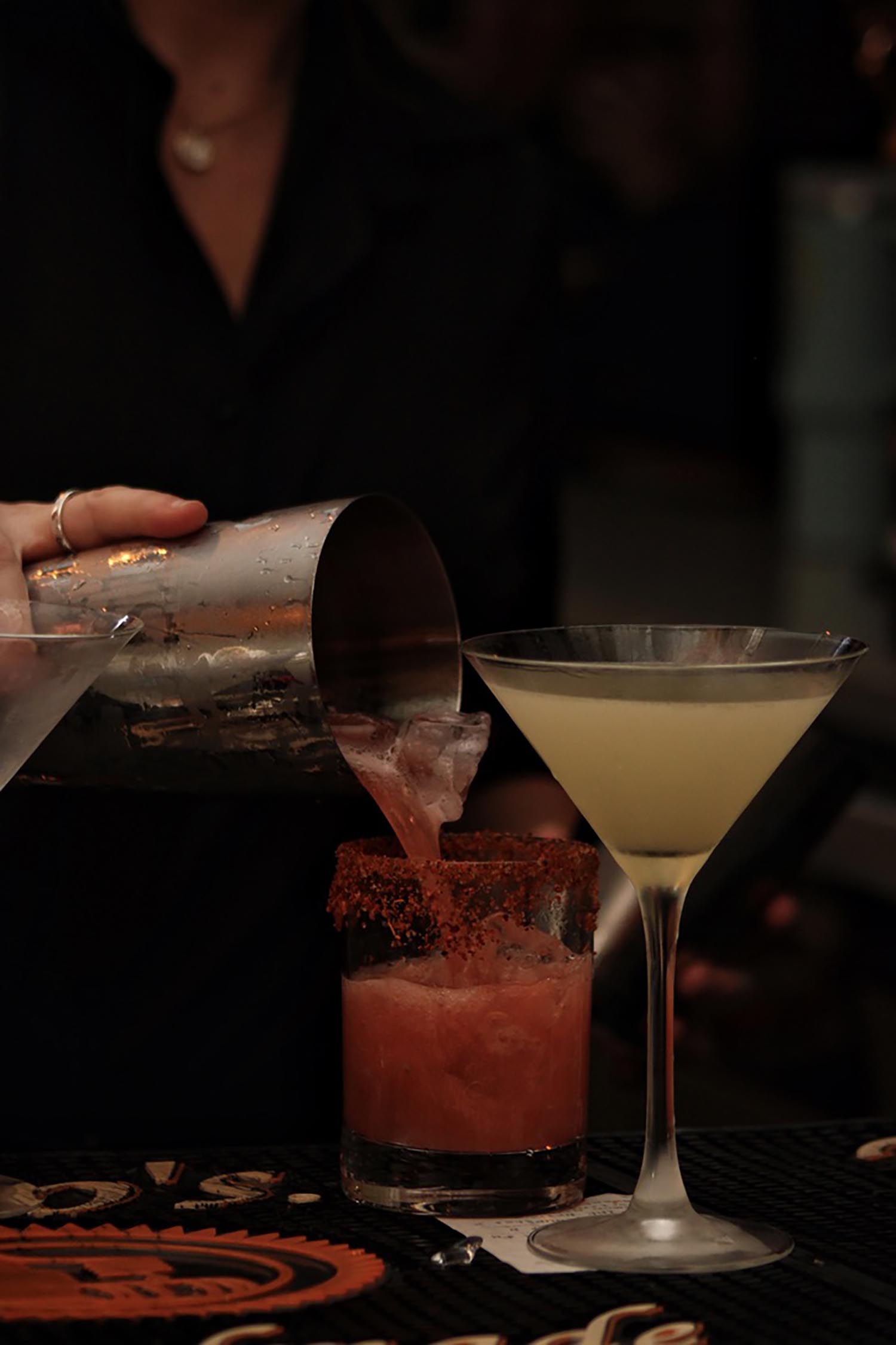 Person pouring a red cocktail with ice into a glass rimmed with chili powder, next to a pale yellow martini glass on a bar counter.